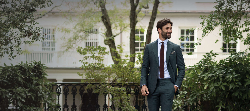 A man outdoors in front of a large ivory house, surrounded by greenery, smiling, and wearing a green suit, burgundy tie, and blue dress shirt.