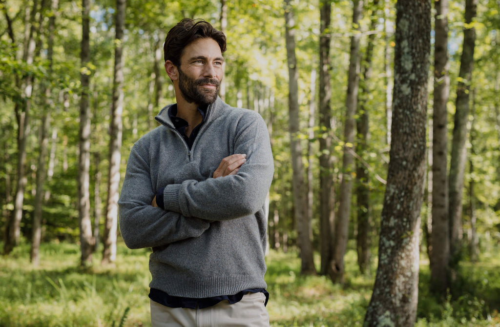 Man standing in a wooded area wearing a light grey soft half zip layering sweater 