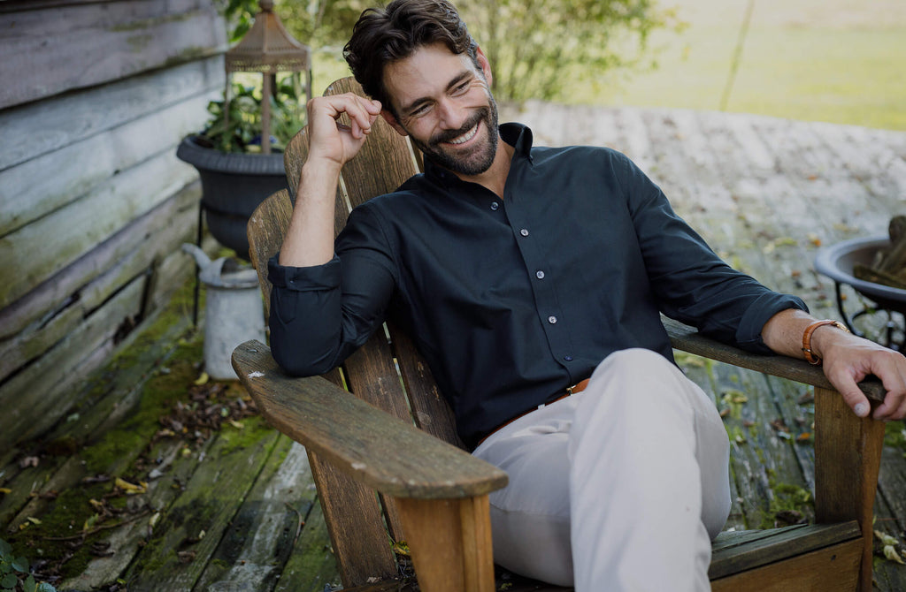 Man smiling sitting on back deck wearing dark green Oxford shirt and tan pants.