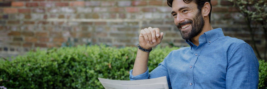 A man smiling wearing the blue Sandoval casual shirt, holding a newspaper, sitting in a garden with a green bush and brick wall behind him.