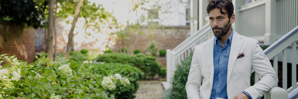A man wearing a blue chambray shirt with an ivory sport coat on top. He's standing in a brick garden courtyard, leaning against a stair railing.
