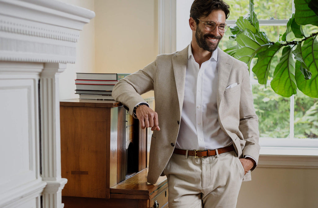 A man leaning against a wooden bookshelf wearing a white dress shirt, ivory sport coat, and tan chinos.