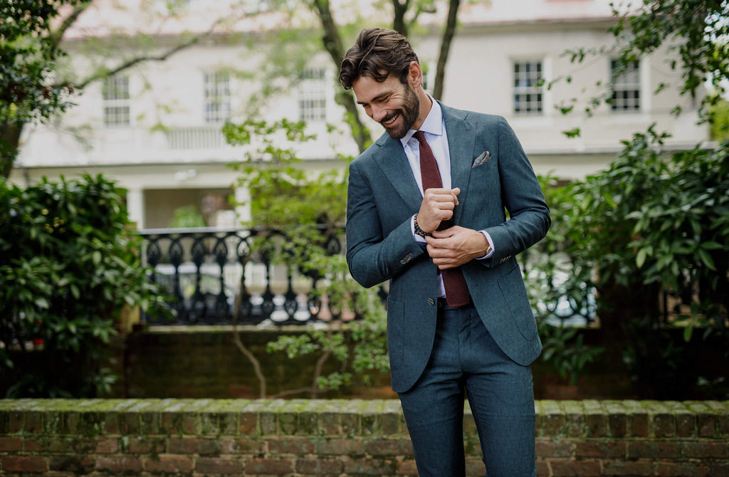 A man outdoors in front of a brick and iron fence wearing a green suit, maroon tie and light blue dress shirt.