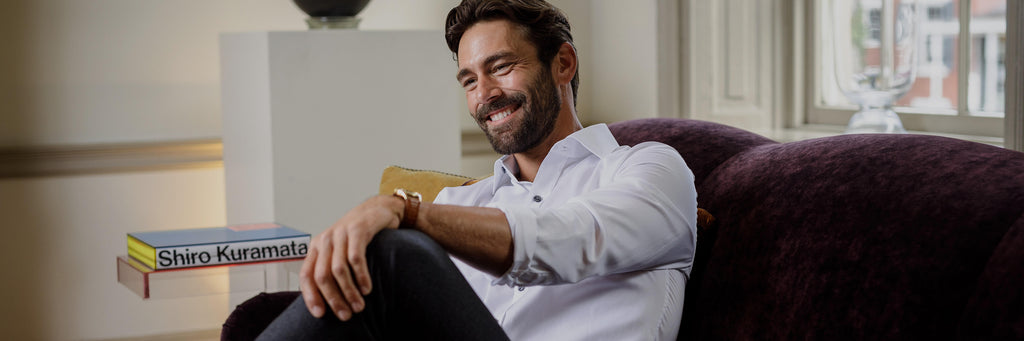 Man smiling while sitting on purple couch wearing the a white dress shirt with smoke buttons.