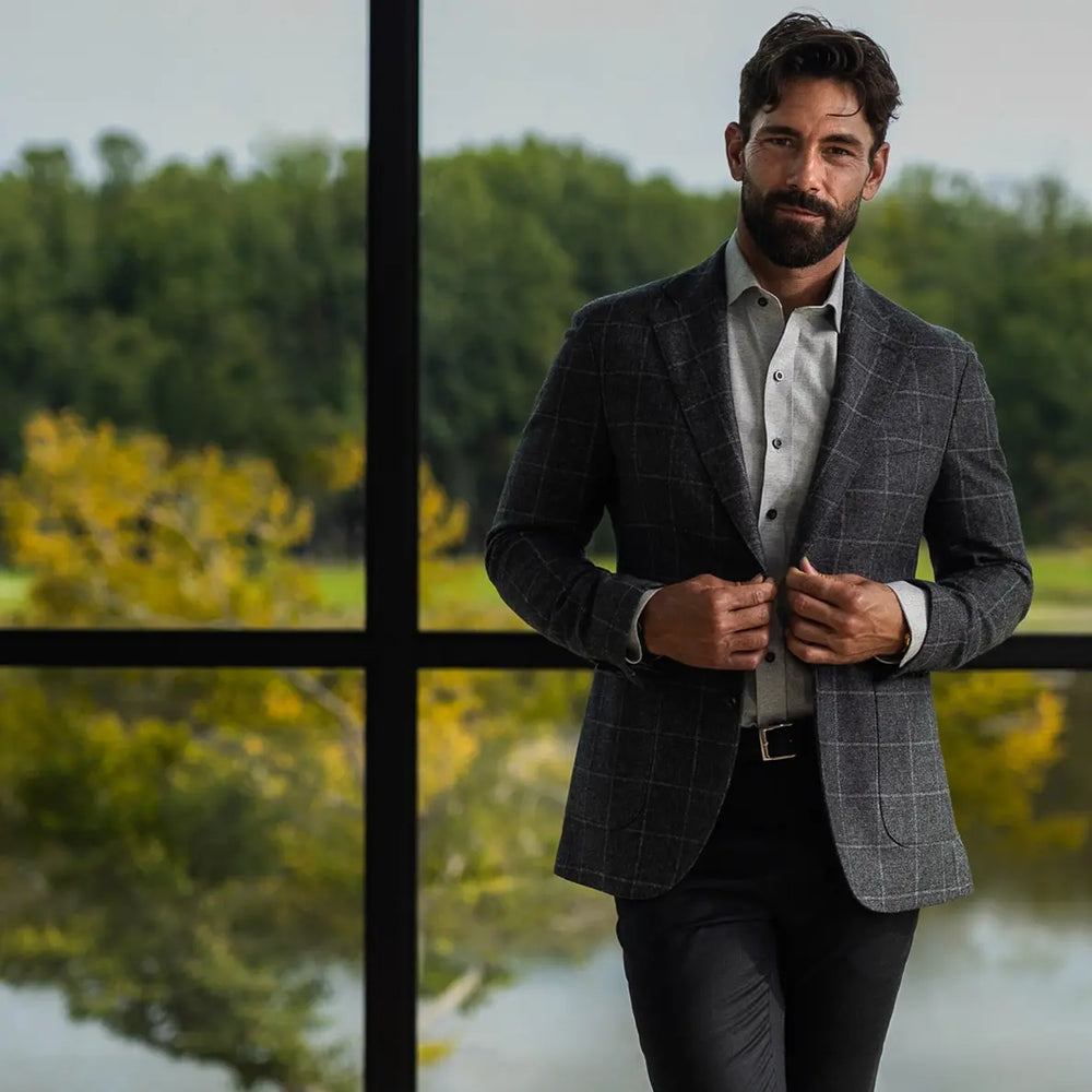 A man wearing a gray windowpane plaid blazer over a light gray button-down shirt and black trousers, standing in front of a large black-framed window with a blurred green and yellow outdoor background.