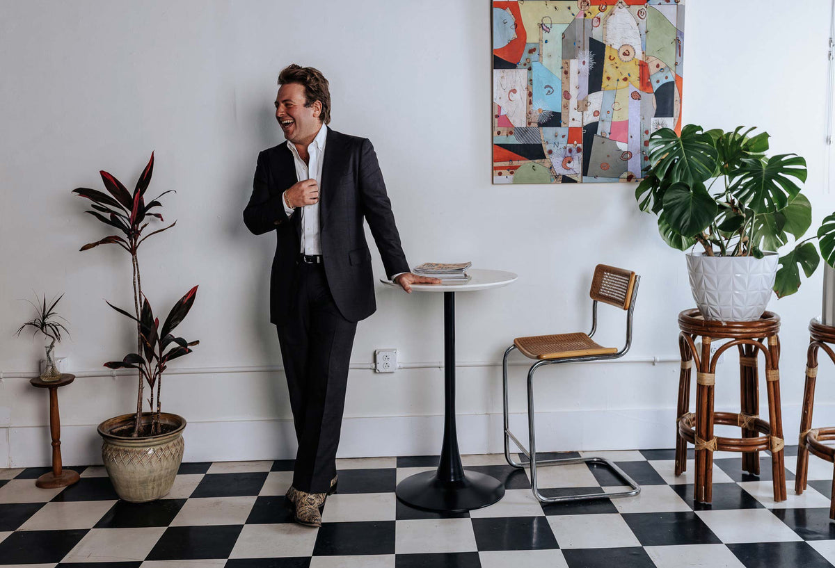 Man in a suit standing in a room with checkered floor, plants, and a table.