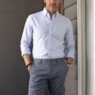 Man wearing a light blue oxford shirt and gray pants standing against a gray wall.