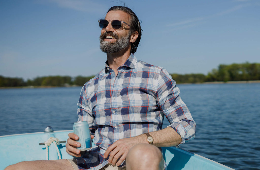 Man sitting on light blue boat in the middle of a lake wearing the Okemo plaid shirt smiling and holding a beer.