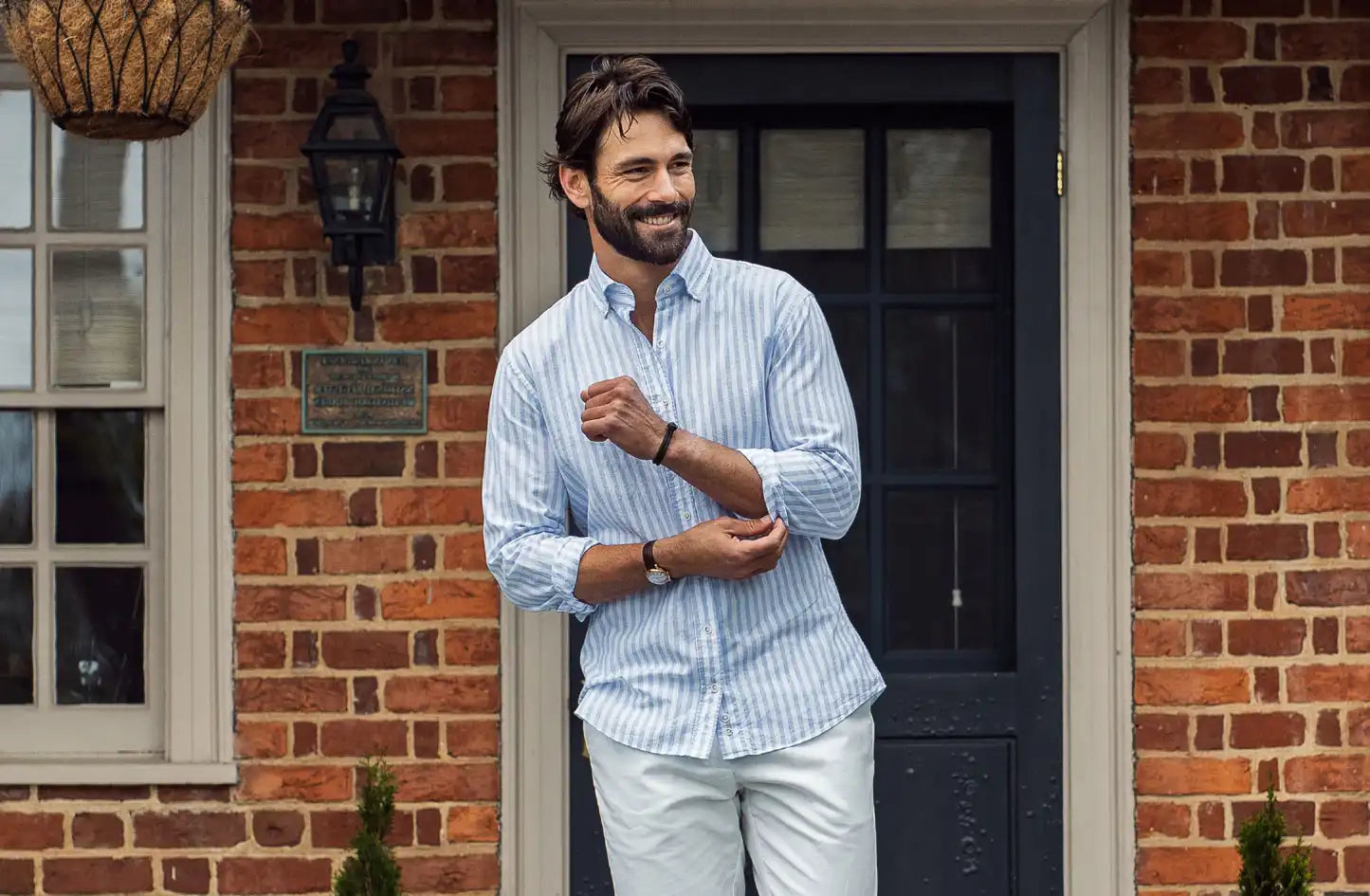 Male model wearing a blue an white striped cotton linen blend shirt by Ledbury. He is standing in front of a brick house with a black doorway. 