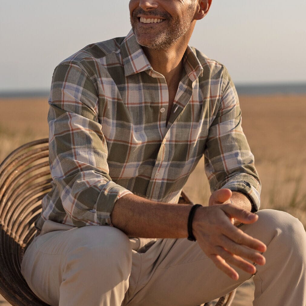 Man sitting outdoors wearing a plaid shirt and beige pants, with a blurred natural background.