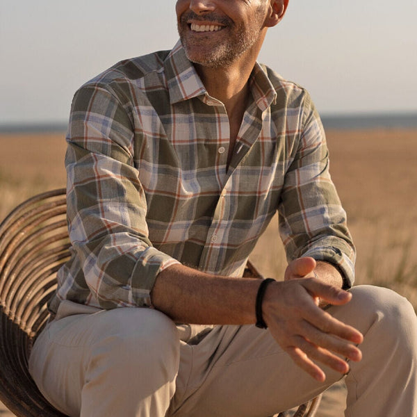 Man sitting outdoors wearing a plaid shirt and beige pants, with a blurred natural background.