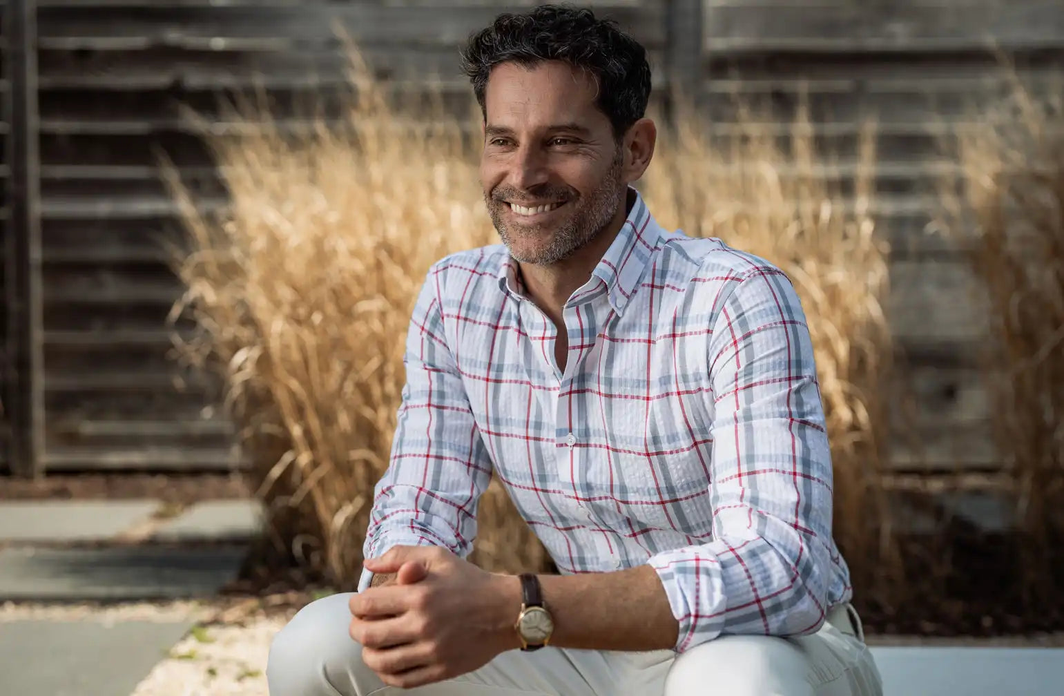 Man wearing a plaid shirt sitting outdoors with dry grass and wooden structure in the background