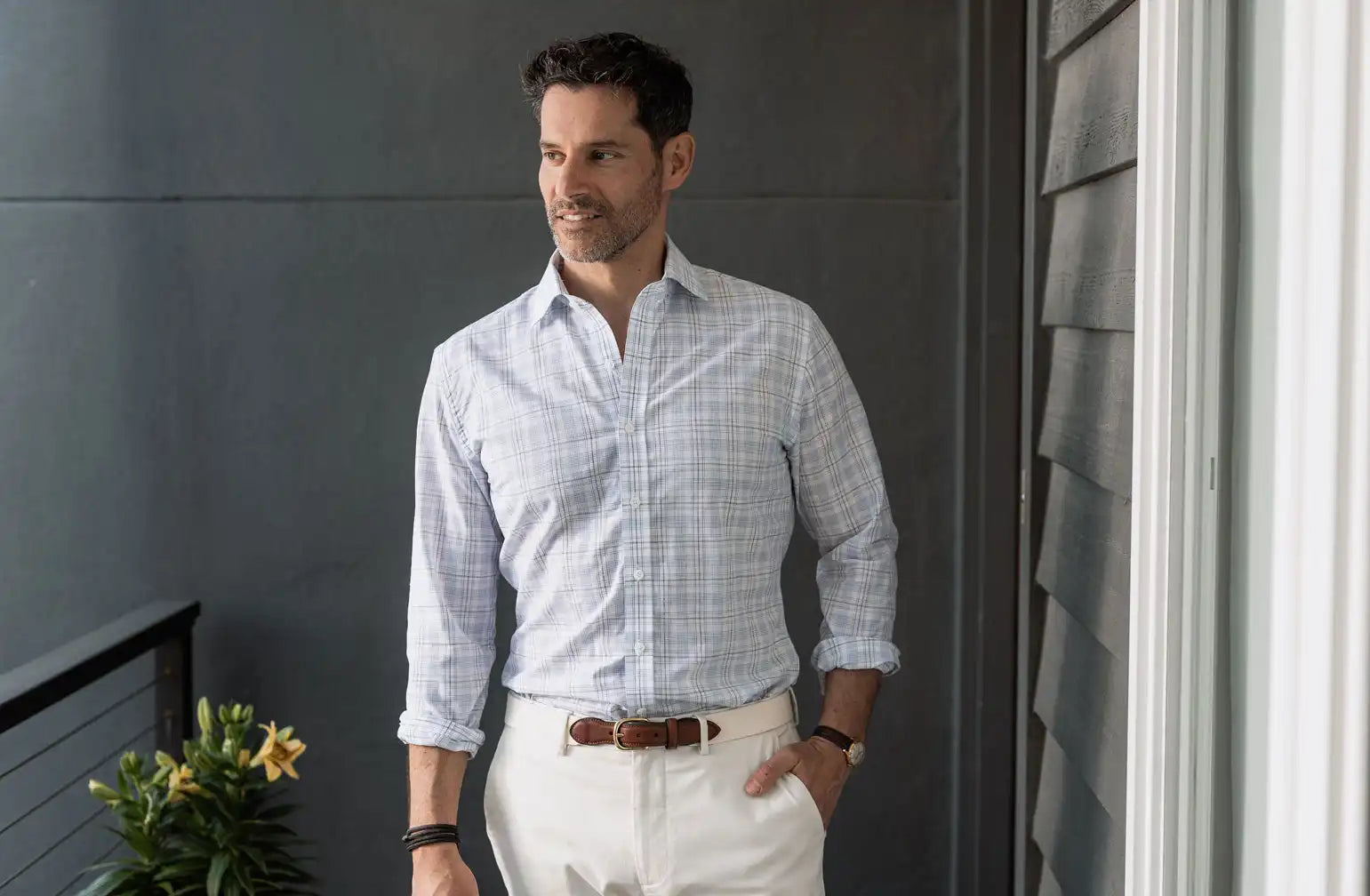 Man wearing a light blue checkered shirt and white pants standing against a gray wall.