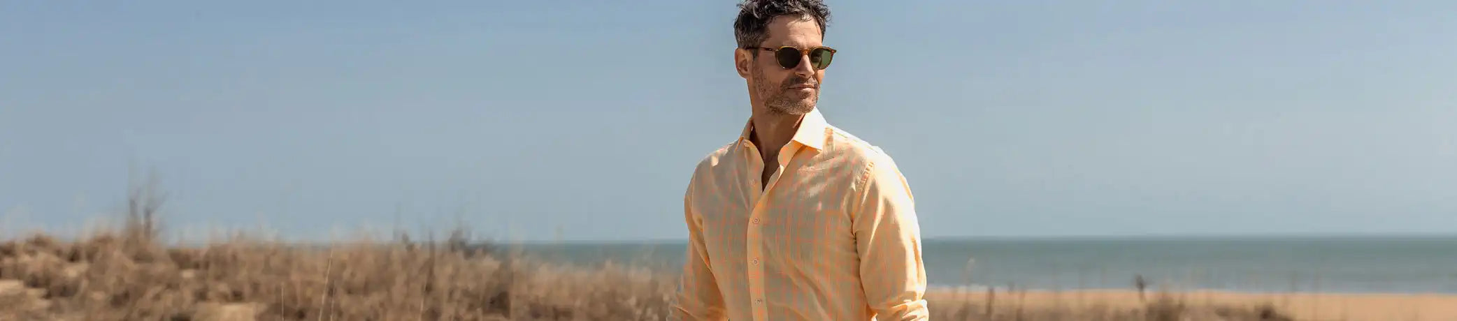 Man wearing sunglasses and a light-colored shirt standing on a beach with clear blue sky.