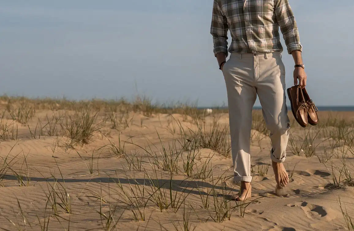 Man walking on a sandy beach with grass, wearing a plaid shirt and light-colored pants.