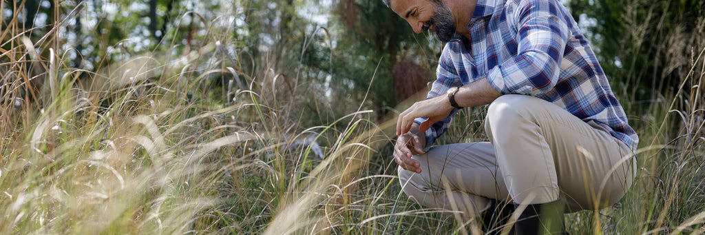 Male model squatting in a field of tall grass wearing tan chinos and a navy and blue plaid summer weight shirt