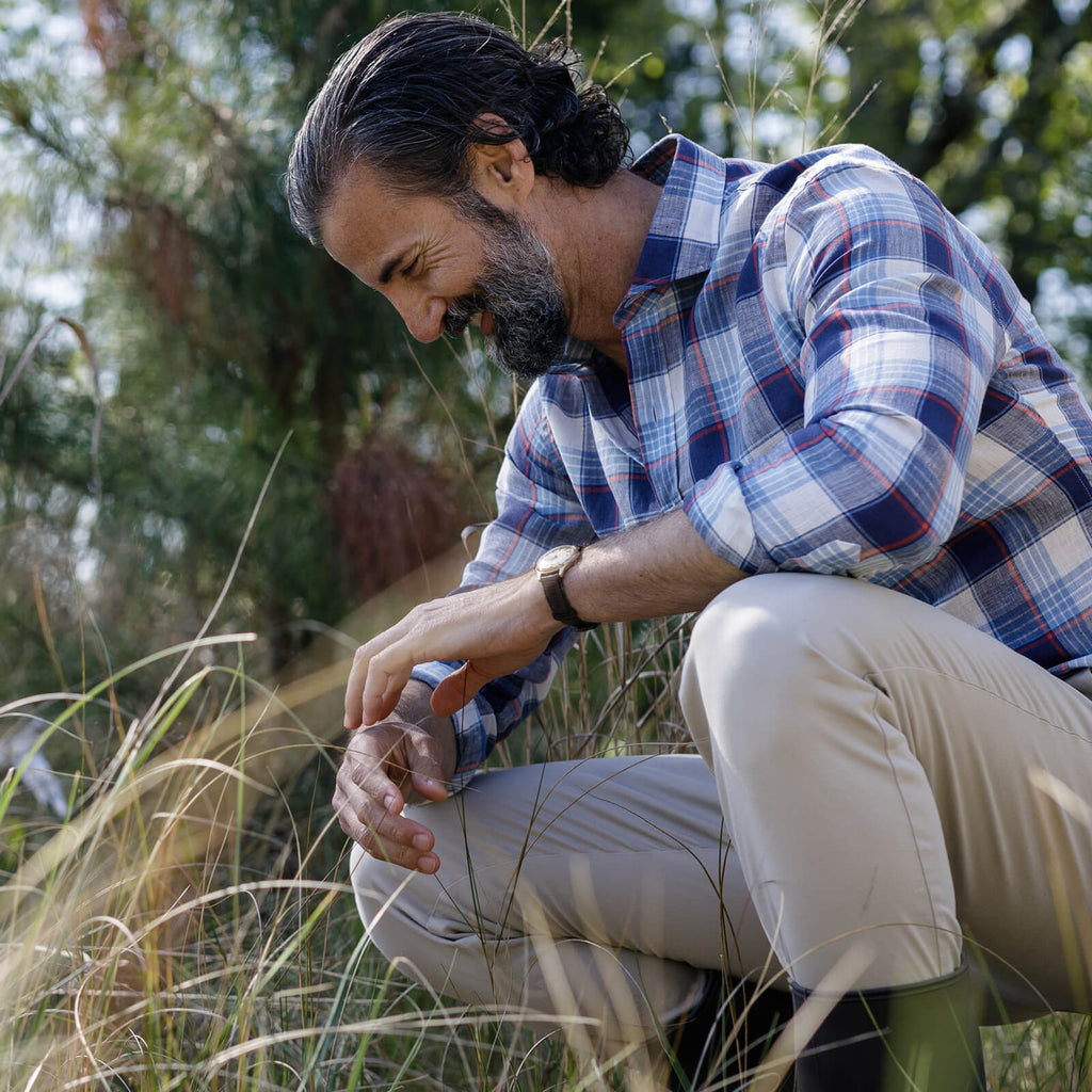 Male model squatting in a field of tall grass wearing tan chinos and a navy and blue plaid summer weight shirt