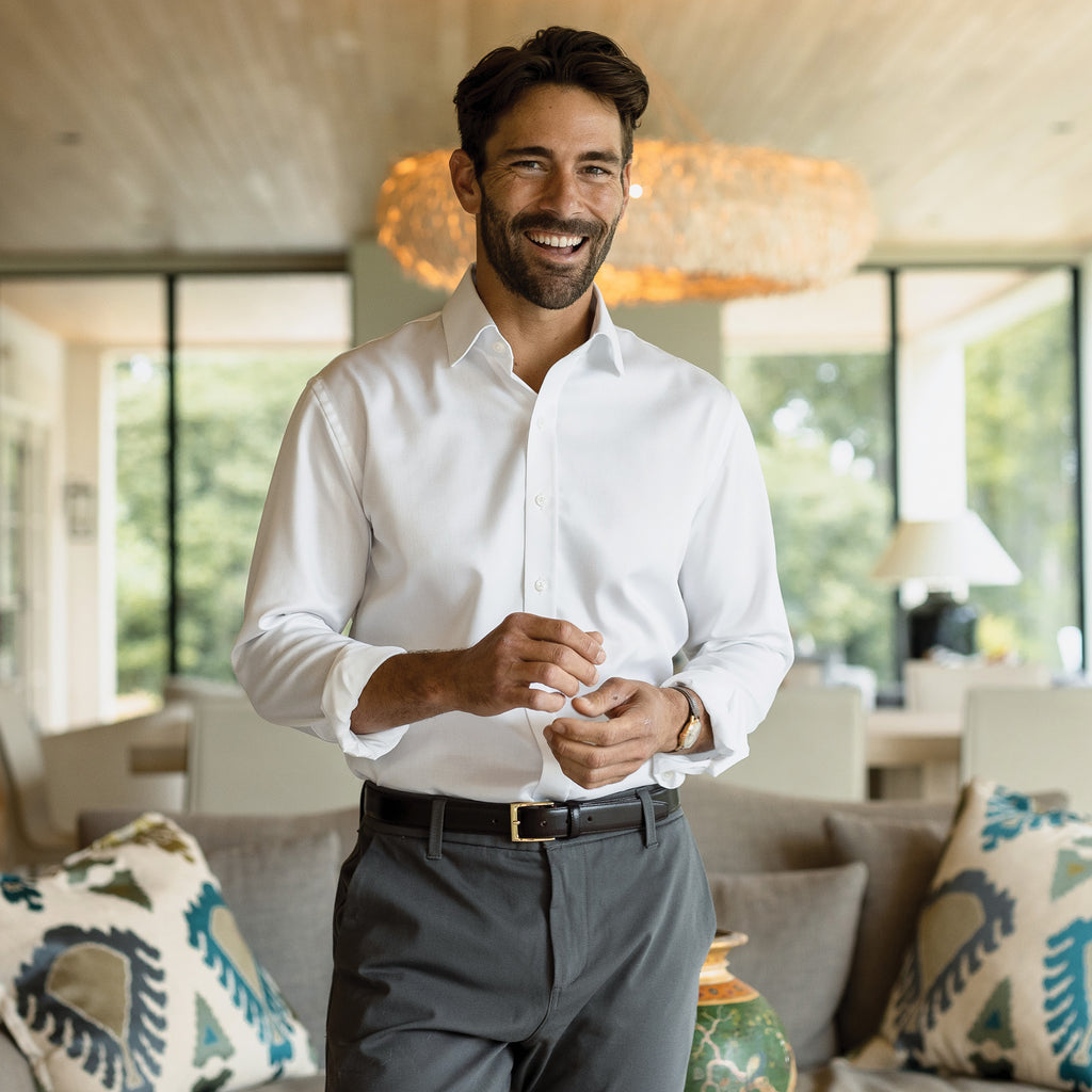 A man smiling inside a home wearing a white dress shirt and gray dress pants.