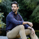 A man sitting outdoors by a body of water wearing a blue and red plaid button-down shirt and tan pants, with a lush green forest background.