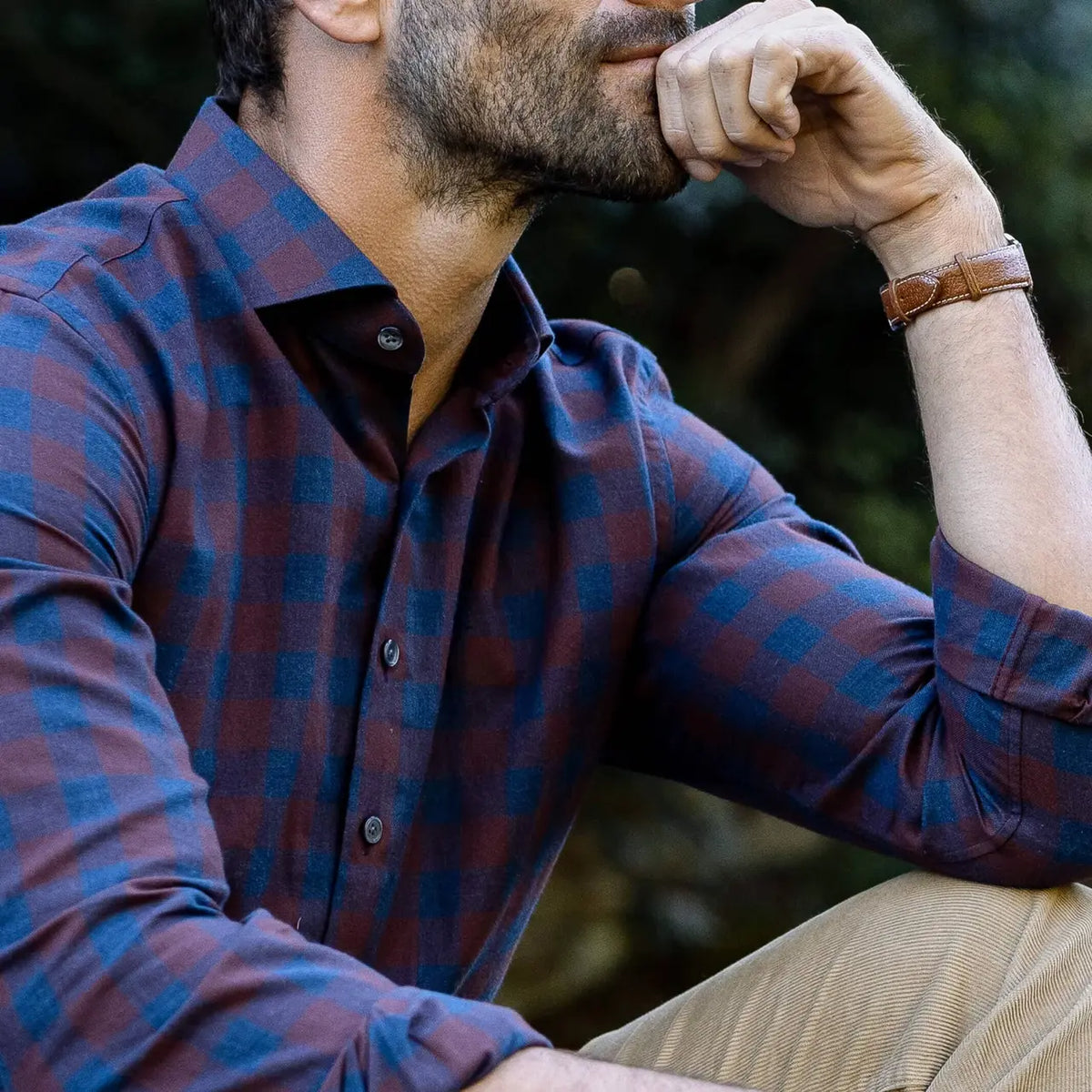 A close-up lifestyle shot of a man wearing a long-sleeve plaid button-down shirt in dark red and blue, paired with khaki pants and a brown leather watch.