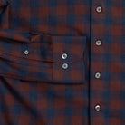 Close-up of a plaid button-up shirt in dark red and navy blue, showing a chest pocket with a flap and smoke grey buttons.