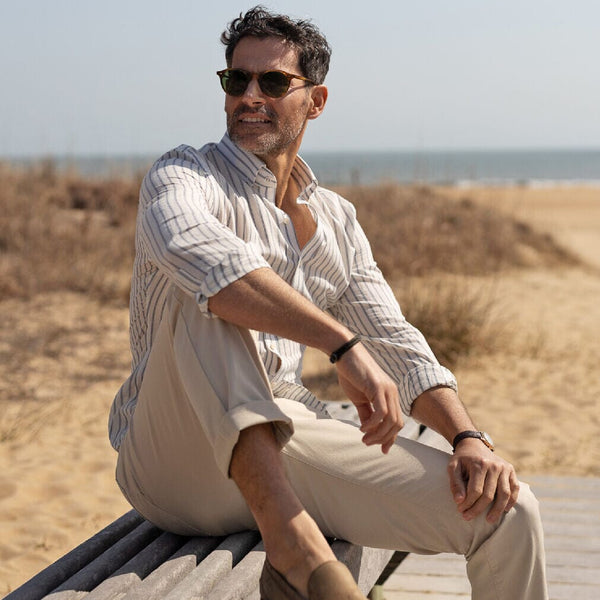 Man sitting on a wooden bench by the beach wearing sunglasses and a light blue striped cotton linen shirt 